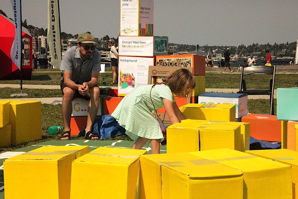 Yellow painted boxes represent housing units. Girl arranges them into a city living space at the Seattle Design Festival Block Party.