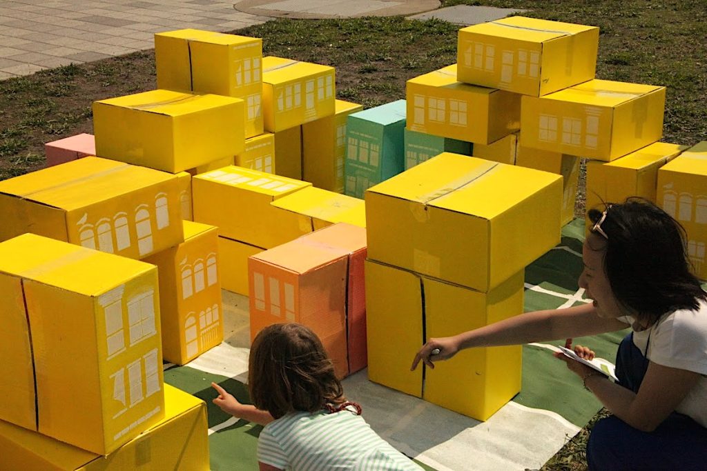 A woman and child point to an arrangement of yellow boxes stacked like a city living space.