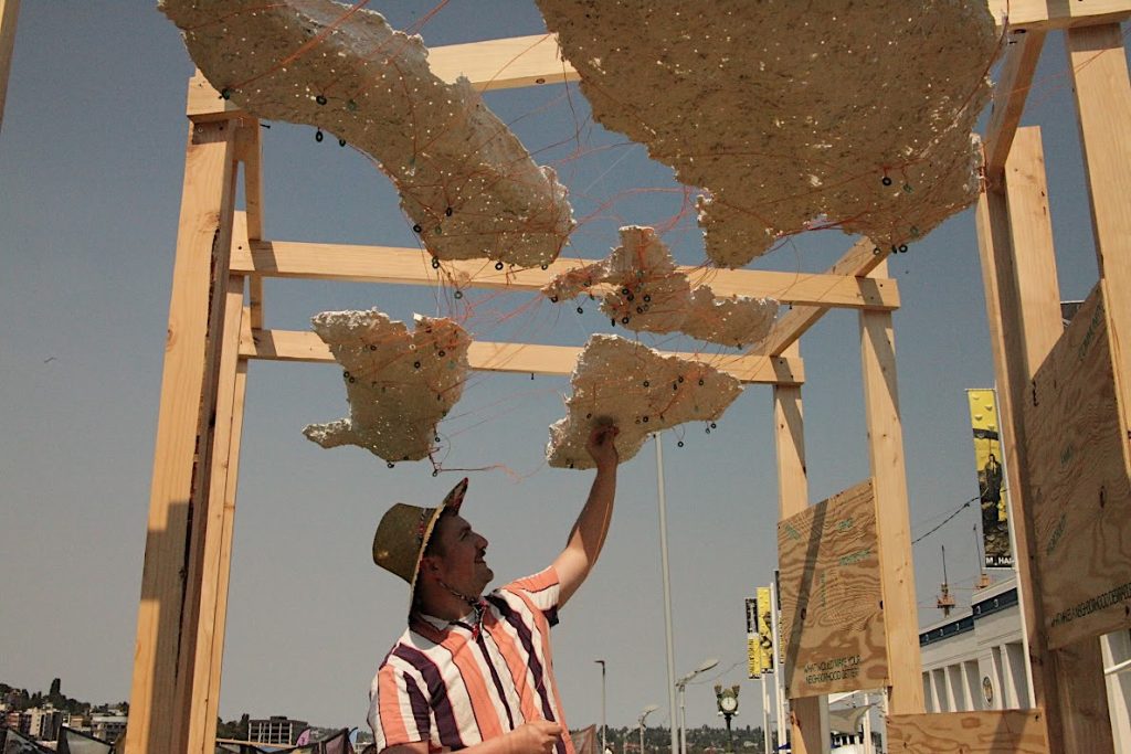 Man with stripped shirt and straw hats moves pieces on strings in a framed cloudlike structure.
