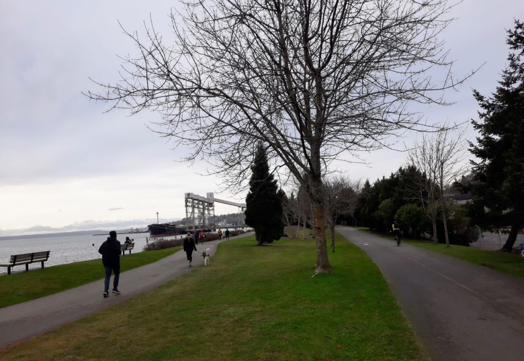 Pedestrian walk on the waterfront trail and a person biking uses the separate multi-use trail to the right.