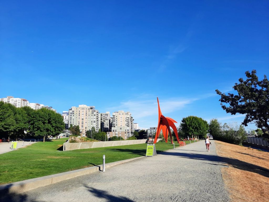 A big red metal Calder sculpture juts out along a wide gravel walking path with midrise Belltown apartments in the background.