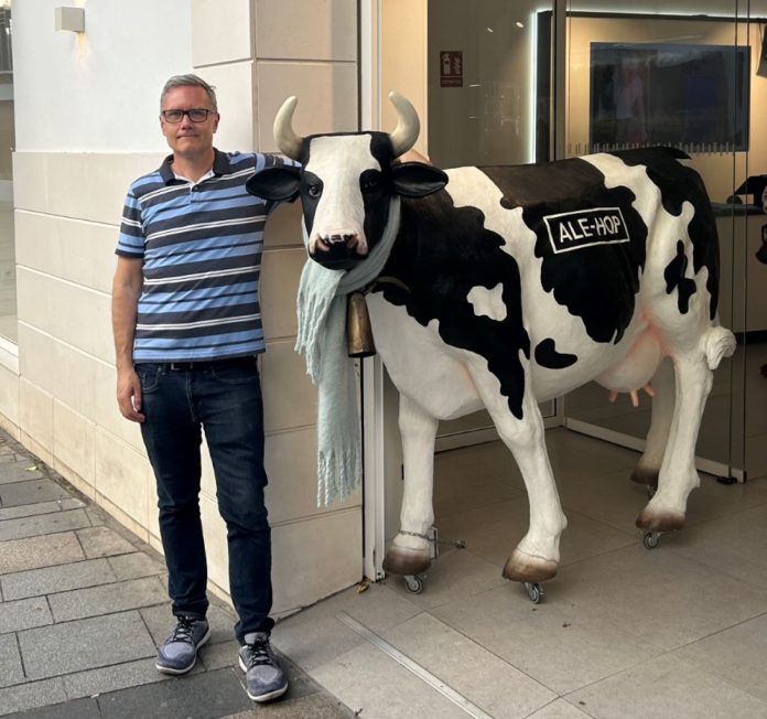 Ray poses with a life-size cow sculpture while traveling in Spain. Ray wears glasses and has sandy-colored hair.