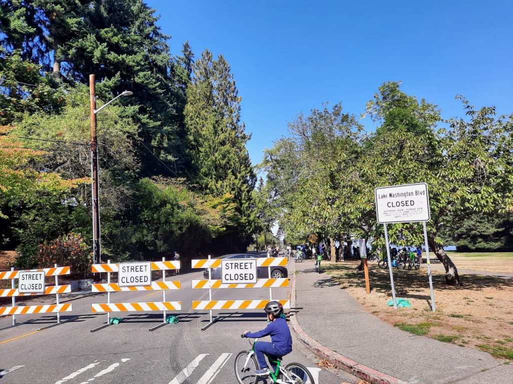Seward Park with Lake Washington Boulevard barricaded to the south. A young kid bikes near the gate with a big group of urbanist bike riders in the background.