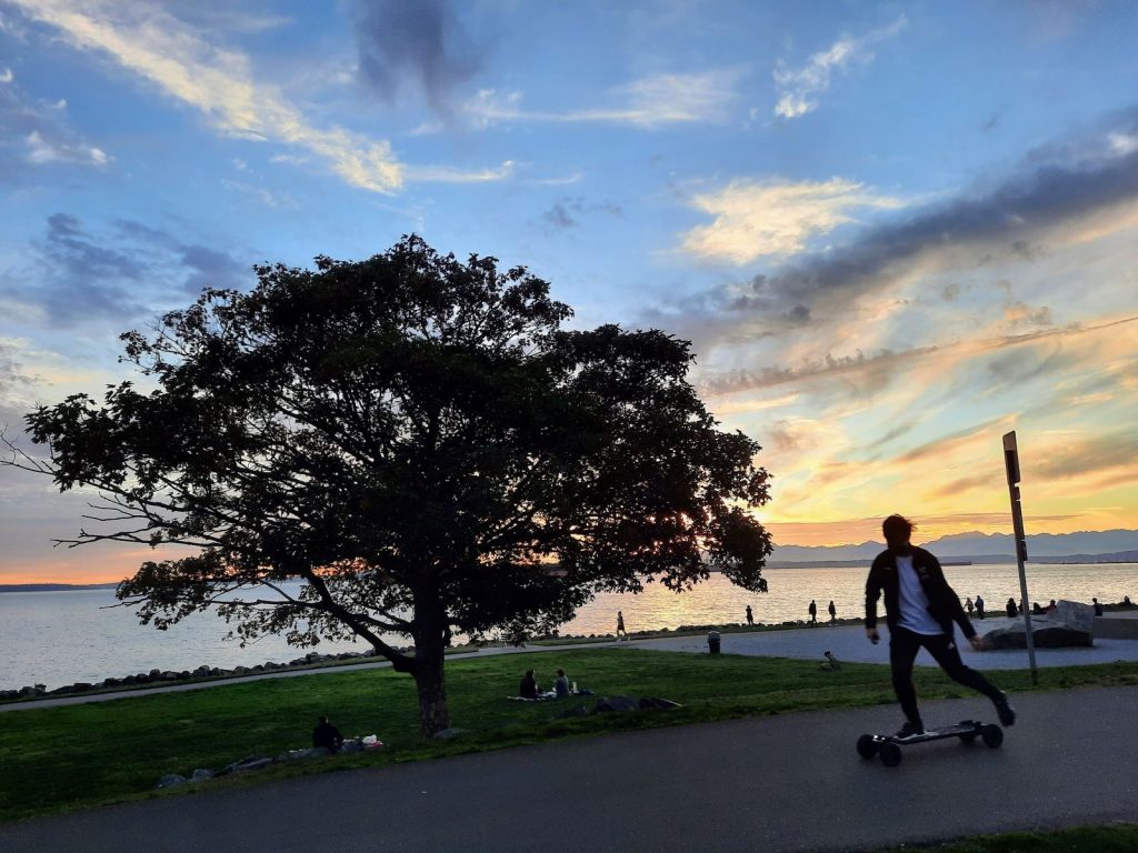 The sun sets in shades of orange over the Olympic Mountains. A wide-canopied tree and a skateboarder are in the foreground and a line of people walking along the waterfront path are in the background.