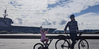 Man on a bicycle with kiddycrank tandem attachment his daughter in pink dress is riding. Behind them is the USS Kitty Hawk aircraft carrier at Sinclair Inlet in Bremerton.