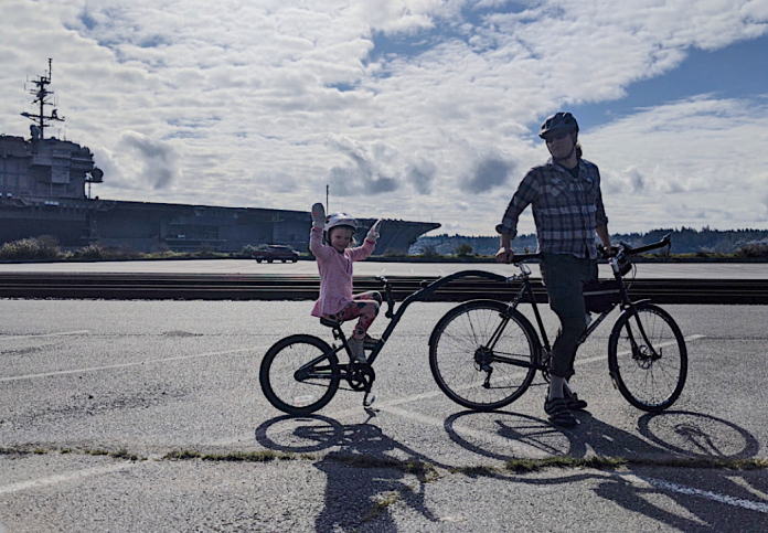 Man on a bicycle with kiddycrank tandem attachment his daughter in pink dress is riding. Behind them is the USS Kitty Hawk aircraft carrier at Sinclair Inlet in Bremerton.