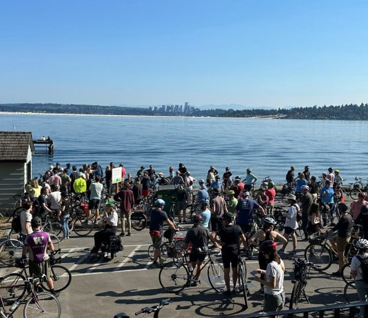 Become a Member, Help Us Close Our Spring Drive Strong A group of about 50 bicyclists stand on the shores of Lake Washington with Bellevue in the background.