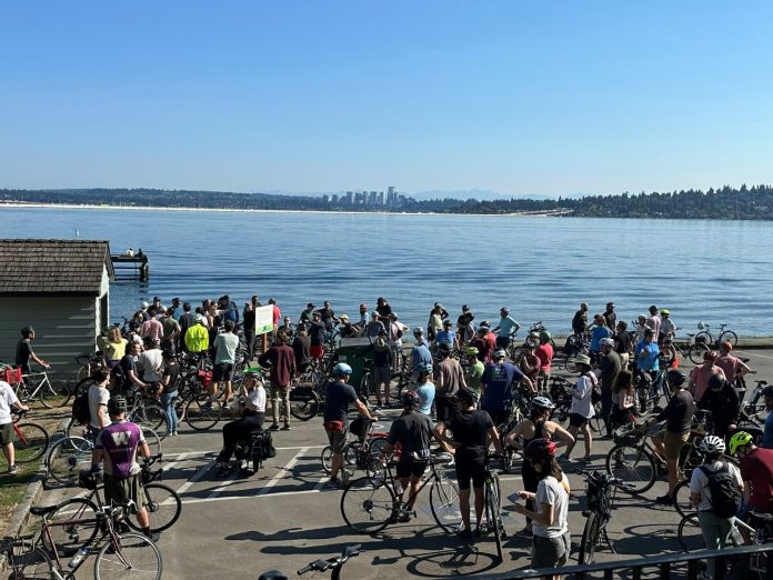 A group of about 50 bicyclists stand on the shores of Lake Washington with Bellevue in the background.