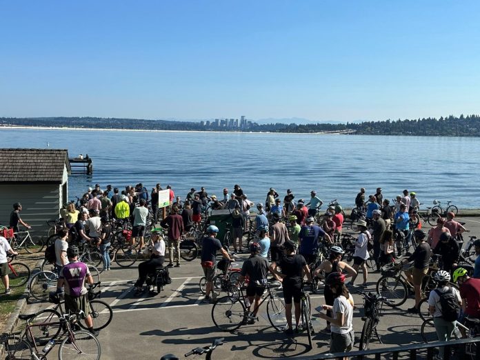 Urbanist-CityNerd-Bike-Ride-at-Mount-Baker-Beach-by-Jackson-Teal-1280 A group of about 50 bicyclists stand on the shores of Lake Washington with Bellevue in the background.