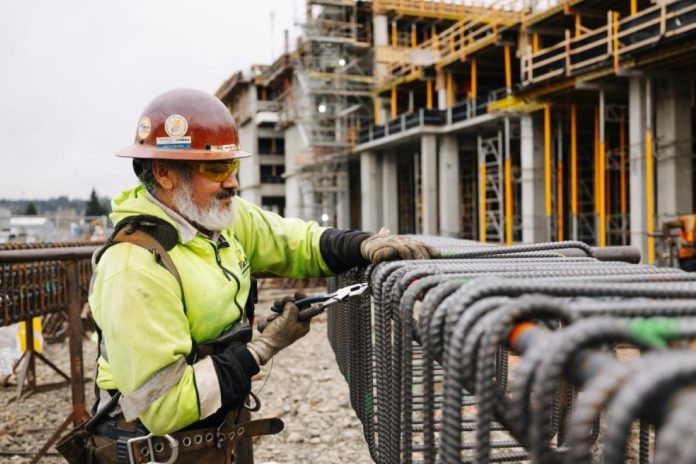 A construction works cuts rebar with the elevated station structure in the background.