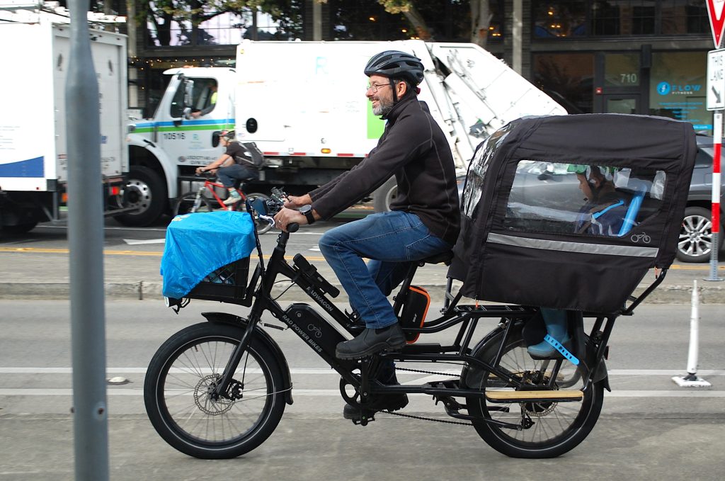 An e-biker with cargo above the front wheel and a child carrier with weather protection over the back wheel. 