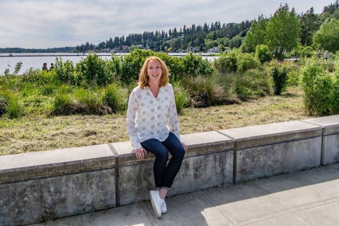 Incumbent candidate Kelli Curtis sitting on a wall with Lake Washington in the background.