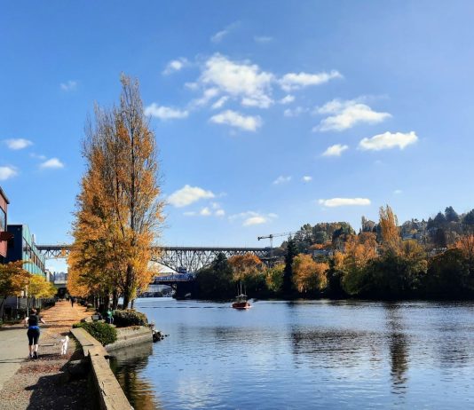 2023 Year-End Update and Fundraising Push People jog and walk along the Burke-Gilman Trail along the canal in Fremont, with a tug boat chuggling along and the bascule bridge and steel high bridge in the background.