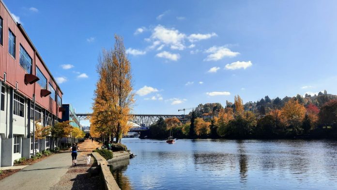 People jog and walk along the Burke-Gilman Trail along the canal in Fremont, with a tug boat chuggling along and the bascule bridge and steel high bridge in the background.