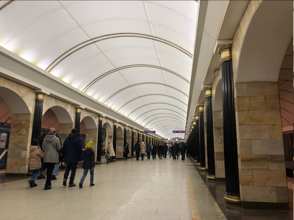 A cylindrical vaulted ceiling brightens up the station with arched arcades along the side. A dozen or so passengers exit the station in small groups.