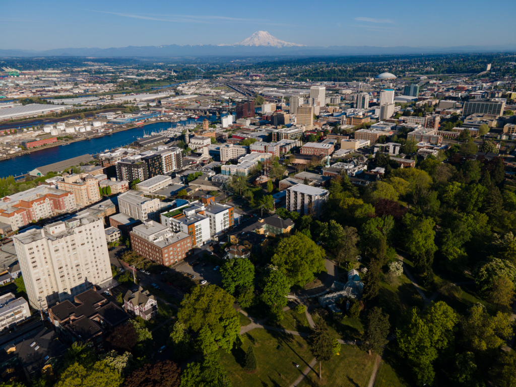 A drone view of downtown Tacoma in late spring with the leafy Wright Park in the foreground and Mount Tahoma (Rainier) in the background. 