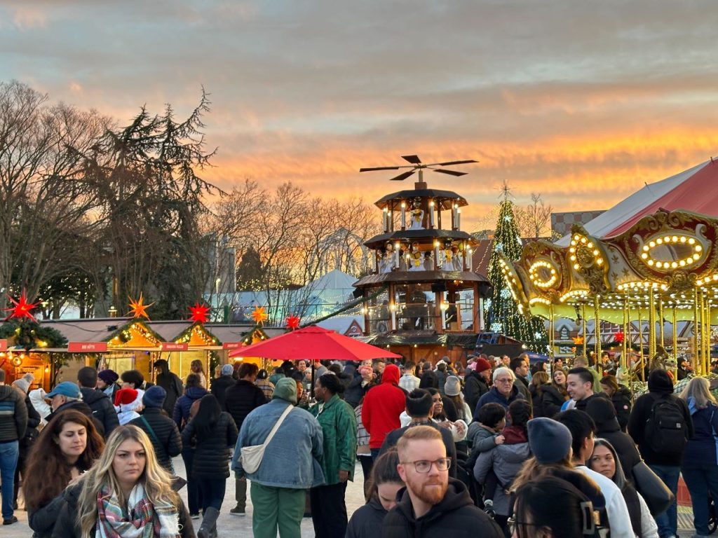 A throng people stand around the pyramid and a merry-go-round. A large Christmas tree and a setting sun is in the background.