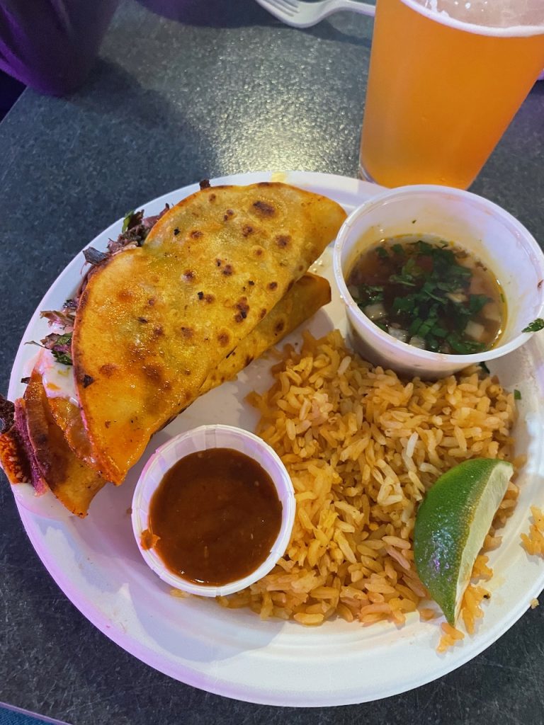 Two tacos with rice and dipping sauce with a remarkably pale beer next to them.