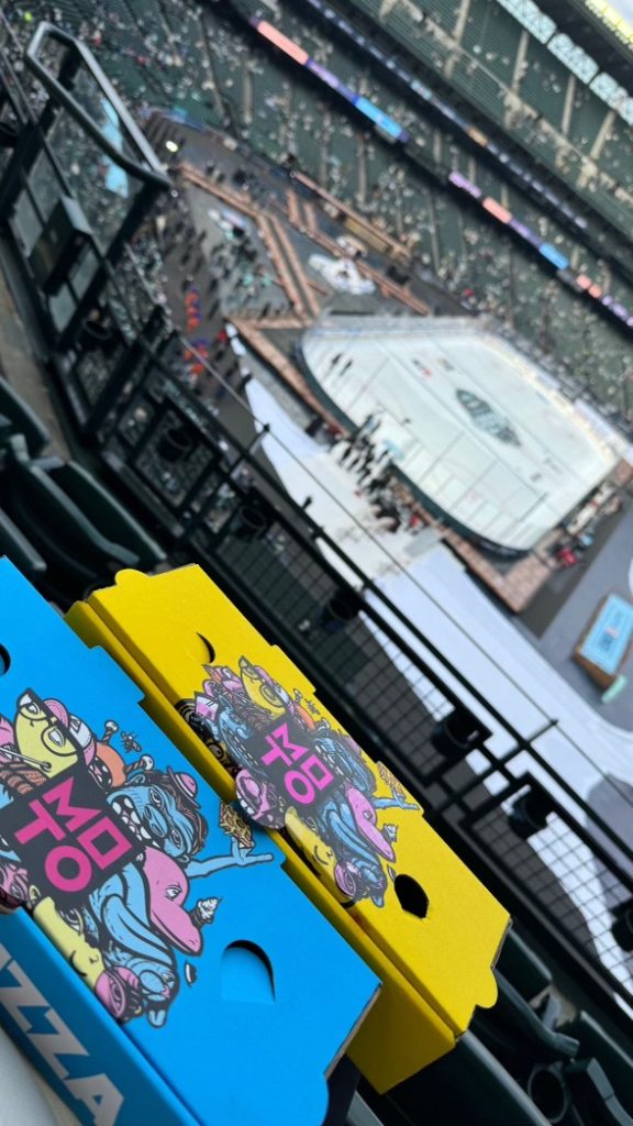 Colorful blue and yellow pizza boxes saying MOTO with view of ice rink on a ball field in the distance.