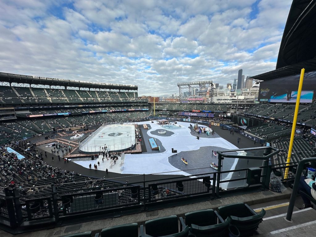 Interior of a baseball stadium with open roof and light clouds, with an ice rink covering the infield and small stages for television and performers in the outfield.