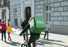 Op-Ed: Gig Worker Minimum Pay Law Makes Seattle More Livable for All A bike delivery person has a large green box with Uber Eats logo on it strapped to their back while riding down an urban streets with lots of pedestrians.