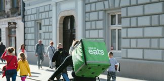 A bike delivery person has a large green box with Uber Eats logo on it strapped to their back while riding down an urban streets with lots of pedestrians.