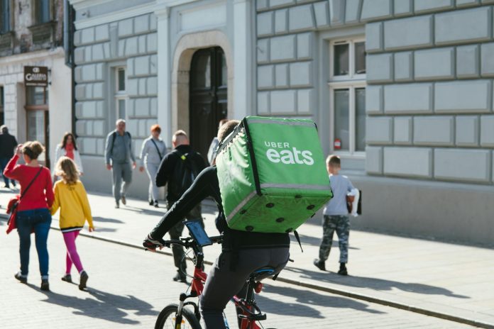A bike delivery person has a large green box with Uber Eats logo on it strapped to their back while riding down an urban streets with lots of pedestrians.