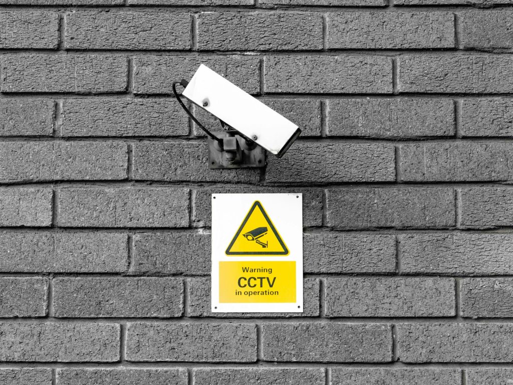 A yellow sign on a brick wall says "Warning CCTV in operation" next to a metal mounted camera.