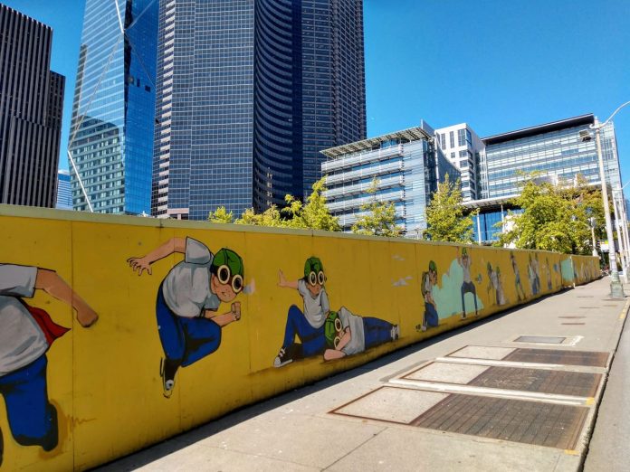 A fence with a pilot boy cartoon figure surrounding Civic Square with Seattle City Hall and the Columbia Tower skyscraper in the background.