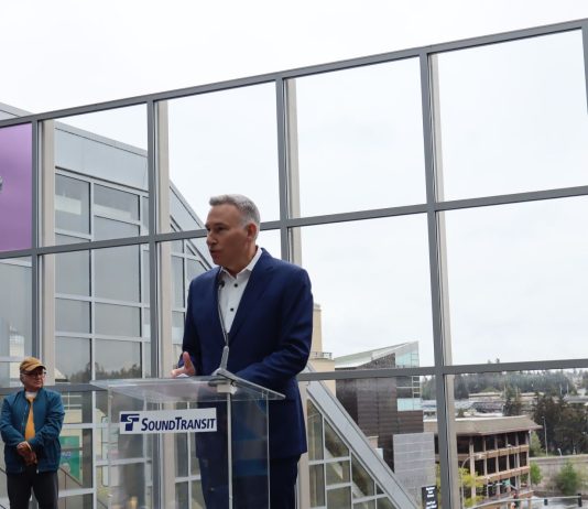 Sound Transit Board Advances Secret ‘Candidate C’ as CEO Pick Constantine wears a suit and stands at a glass lectern in from of the Downtown Bellevue Station plaza overlook of the station platform below.