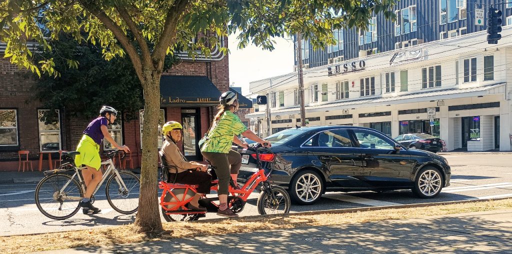 Two set of bikes ride alongside a car with one bike carrying a seated rider in tow.