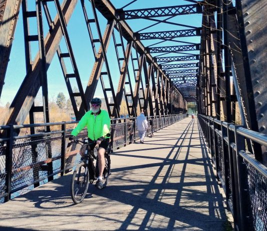 Washington’s E-bike Rebate Program Launches Wednesday A person bikes on a trestle bridge with a few pedestrians in the background.