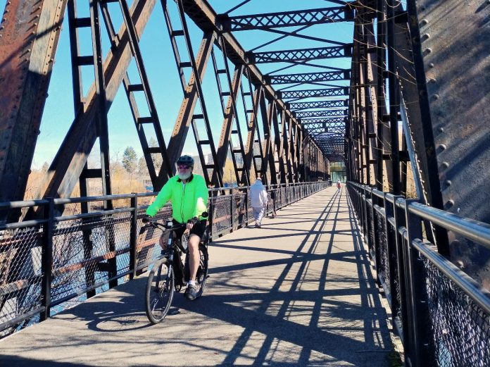 A person bikes on a trestle bridge with a few pedestrians in the background.