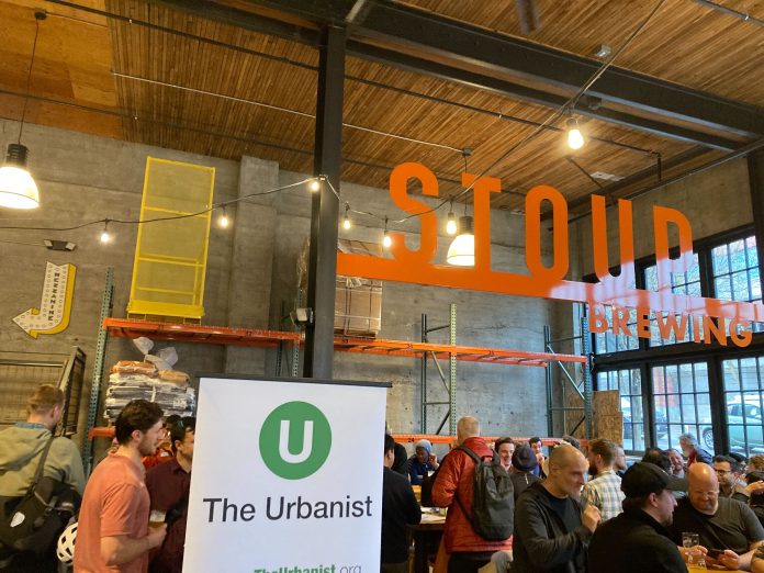 An Urbanist banner standing in a corner of a brewery with a few dozen people gathered chatting.
