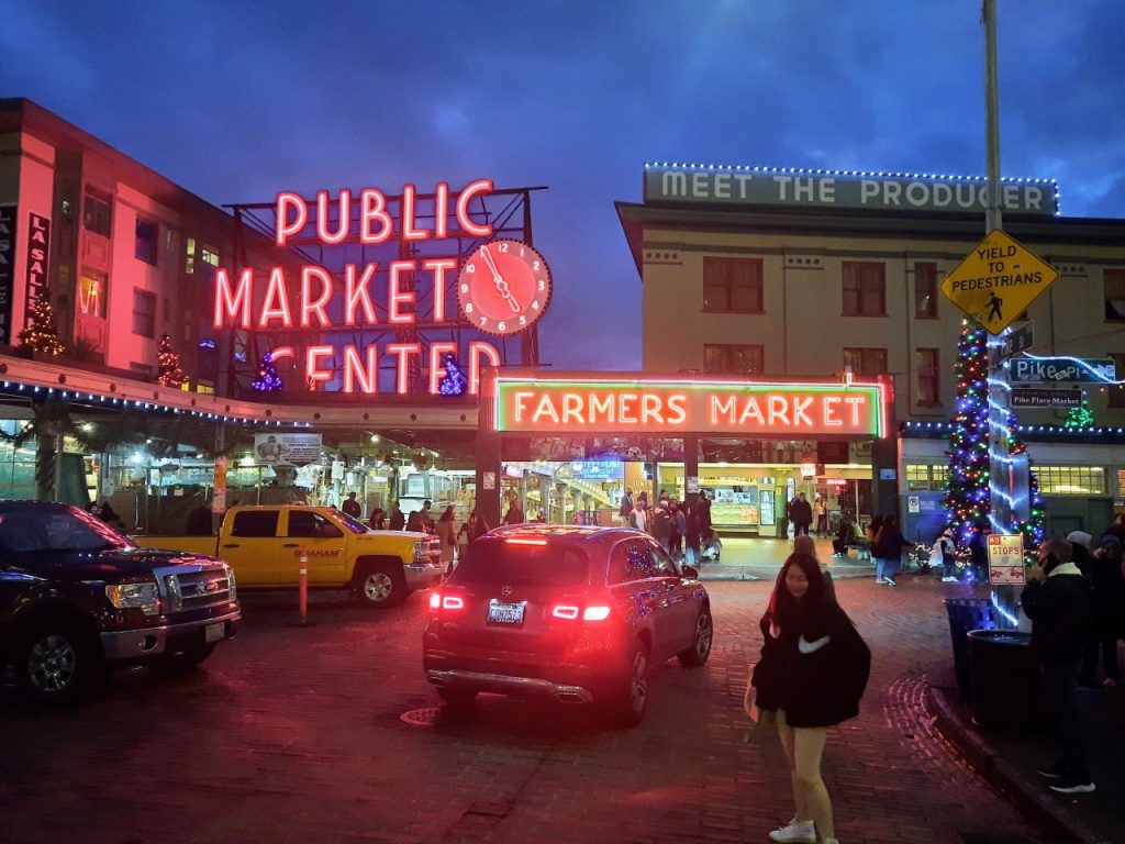 A car turns onto Pike Place next to a pedestrian. The neon Pike Place Market sign pops at dusk.