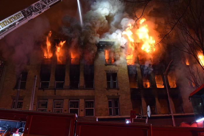 Fire and smoke pours out of upper story windows of an old brick building with a fire crane seeking to control them.