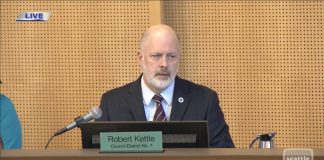 Kattle wears a blue suit and light gray beard and sits on Council dais.