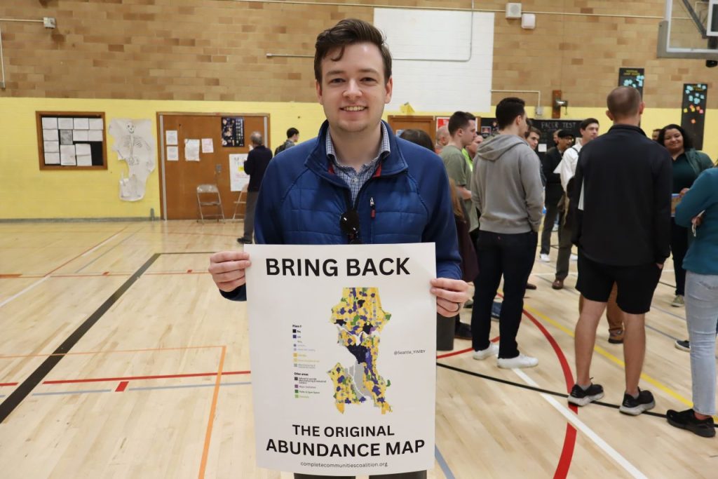 Erik stands in a gymnasium with a sign reading "Bring back the original abundance map."