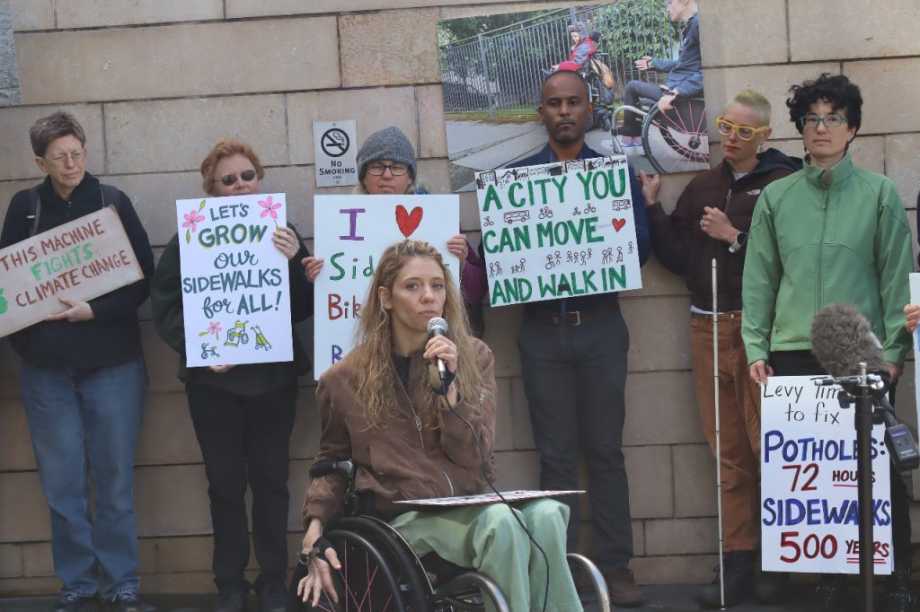 Cecilia hold a microphone and is in a wheelchair. Behind her fellow advocates hold signs saying "This machine fights climate change" with a picture of a bike, "Let's grow our sidewalks for all. "A city you can move and walk in." "Levy time to fix potholes: 72 hours. Sidewalks 500 years."