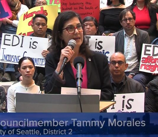 Rivera Walks Back Raid of Anti-Displacement Funds, Claims Critics Are Misinformed Morales speaks at a podium and several rows of supporters hold signs in support of the EDI program and against the budget raid.