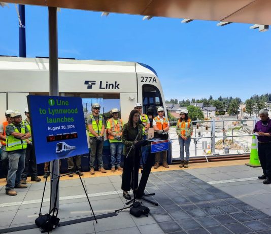 Sound Transit Begins Lynnwood Link Testing, Countdown Clock Mestas stands next to a countdown clock with construction workers lined up behind her in front of a Link train.