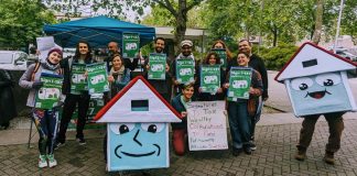 A group of about 20 advocates pose with signs next to House-y the social housing mascot