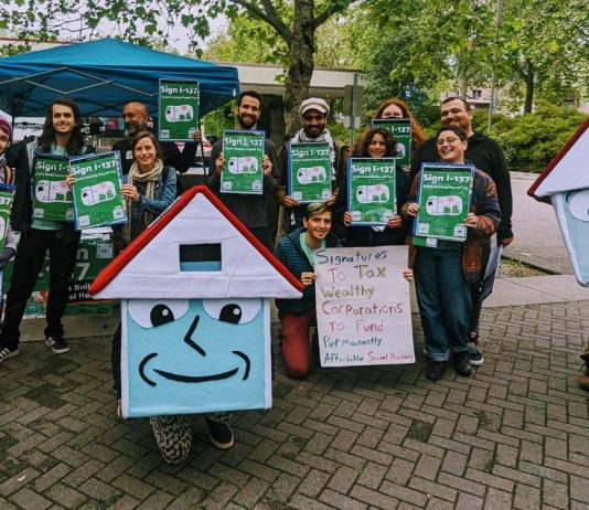 Harrell Proposes $2 Million Loan to Kickstart Seattle Social Housing Developer A group of about 20 advocates pose with signs next to House-y the social housing mascot