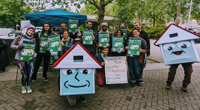 Seattle Social Housing Staffs Up, Nets $115 Million A group of about 20 advocates pose with signs next to House-y the social housing mascot