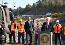 WSDOT Head Millar Wants $150 Million Annually to Fix Washington’s ‘Stroads’ Millar wears a suit and stands at a lectern with Governor Inslee and a group of orange vest wearing construction workers standing behind him. A busy freeway is in the background.
