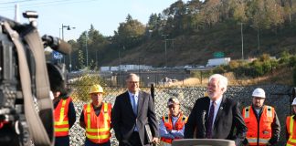 WSDOT Head Millar Wants $150 Million Annually to Fix Washington’s ‘Stroads’ Millar wears a suit and stands at a lectern with Governor Inslee and a group of orange vest wearing construction workers standing behind him. A busy freeway is in the background.