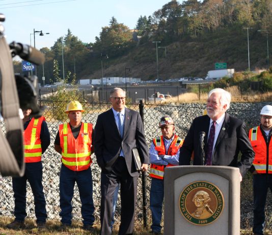WSDOT Head Millar Wants $150 Million Annually to Fix Washington’s ‘Stroads’ Millar wears a suit and stands at a lectern with Governor Inslee and a group of orange vest wearing construction workers standing behind him. A busy freeway is in the background.