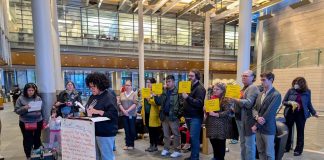 Amid Budget Showdown, Advocates Mount Defense of JumpStart Housing Funds A sign reads Resilient communities on the lectern in the lobby of city hall and people stand behind the speaker holding signs saying a protest JumpStart commitments and invest on Seattle's future.
