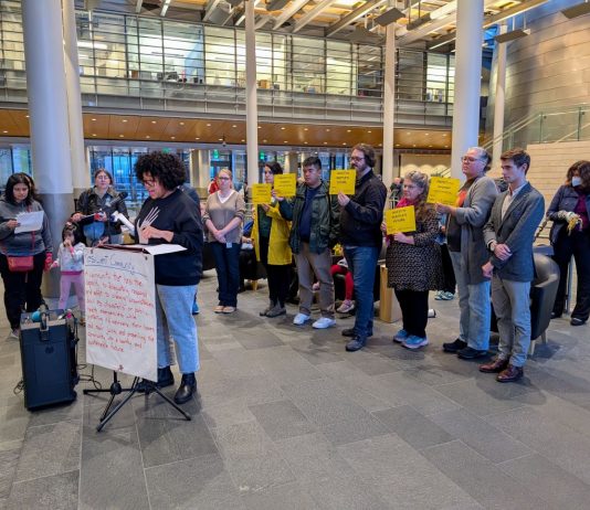 Amid Budget Showdown, Advocates Mount Defense of JumpStart Housing Funds A sign reads Resilient communities on the lectern in the lobby of city hall and people stand behind the speaker holding signs saying a protest JumpStart commitments and invest on Seattle's future.