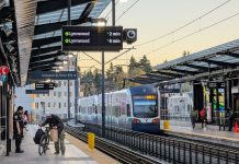 Op-Ed: State Must Reform Sound Transit to Keep Expansion on Track A southbound train departs with an apartment building in the background. A man rolls a bike on the platform and security guards stand around.